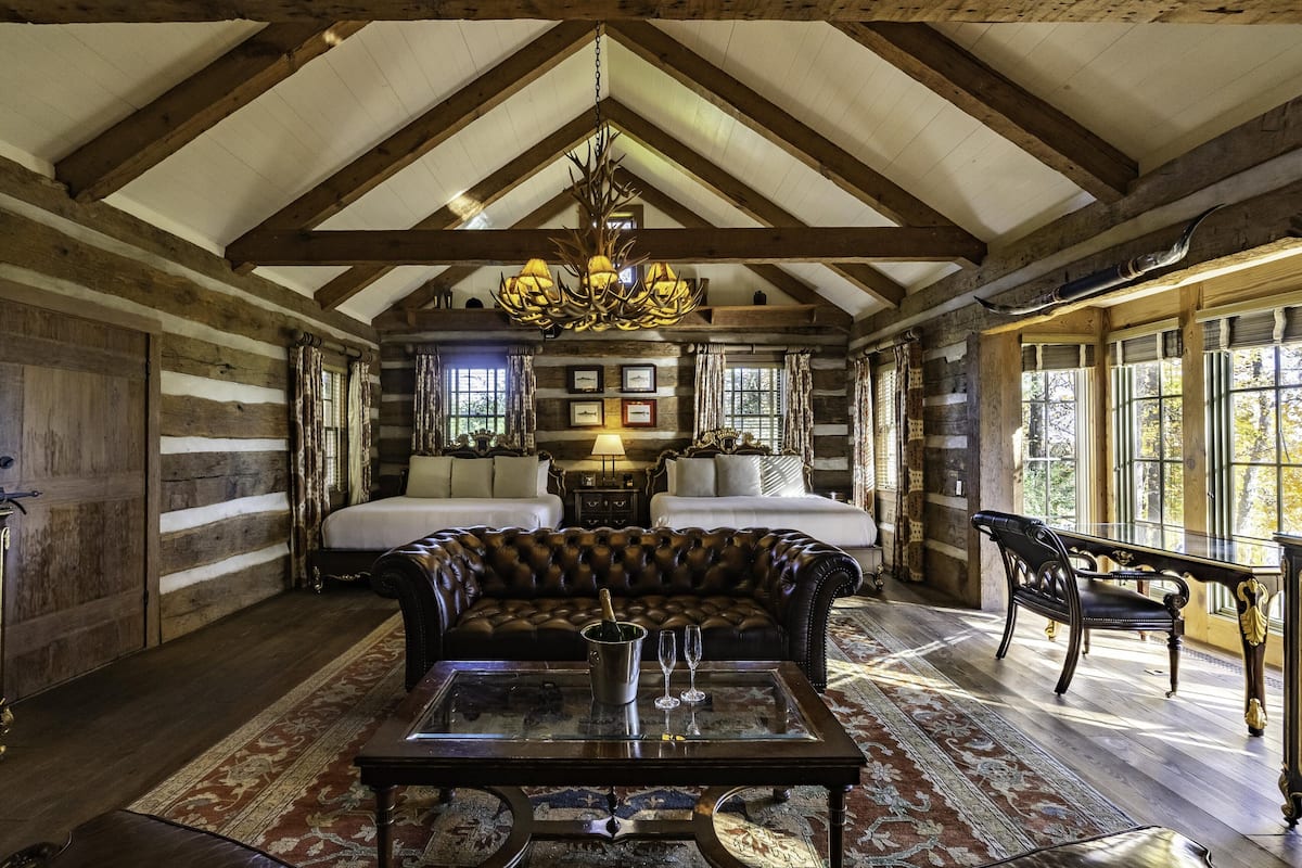 A log cabin interior with a large bed, a leather sofa, a dining table, and a chandelier.
