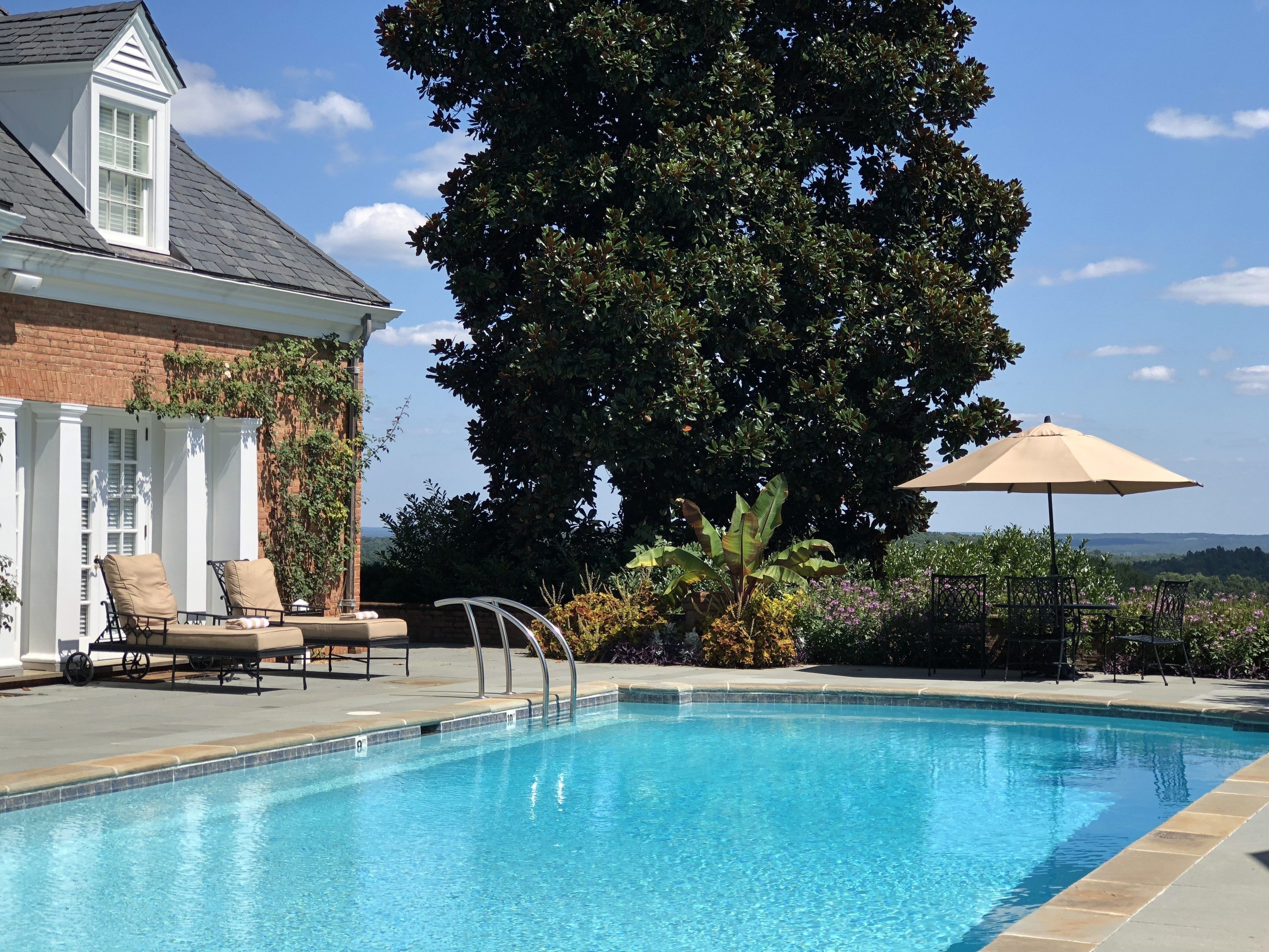 A swimming pool with lounge chairs and an umbrella, surrounded by greenery and a brick building.