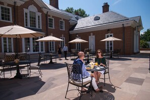 An outdoor patio with tables and chairs, a brick building in the background, and a statue in the center. - Albemarle Estate at Trump Winery (Charlottesville)