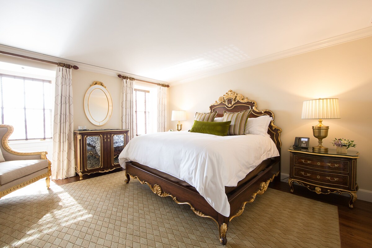 A spacious bedroom with a large bed, a nightstand with a lamp, a dresser, and a chair.