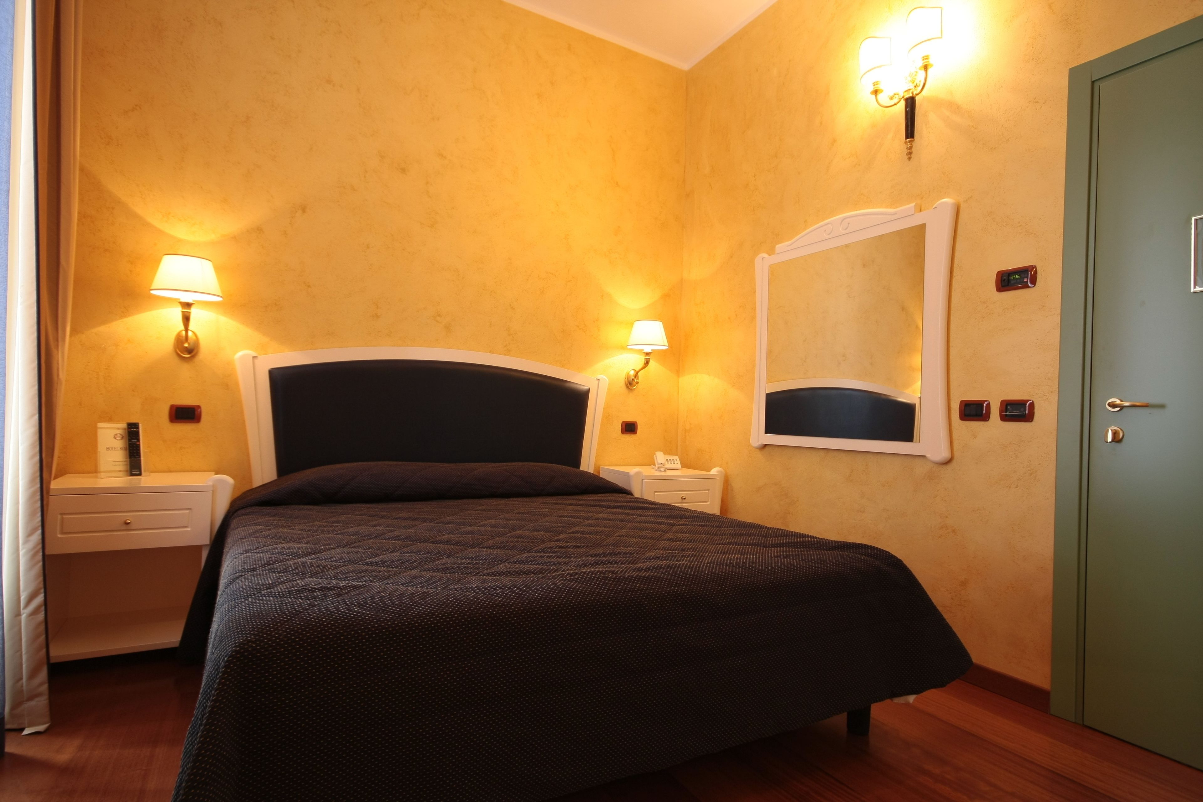 Habitación de hotel con una cama grande, dos mesitas de noche, una mesita auxiliar, una puerta verde y lámparas fijadas a la pared.