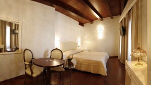 Un dormitorio con cama, mesita de noche, silla, una mesa redonda con dos copas de vino, espejo y lámpara.