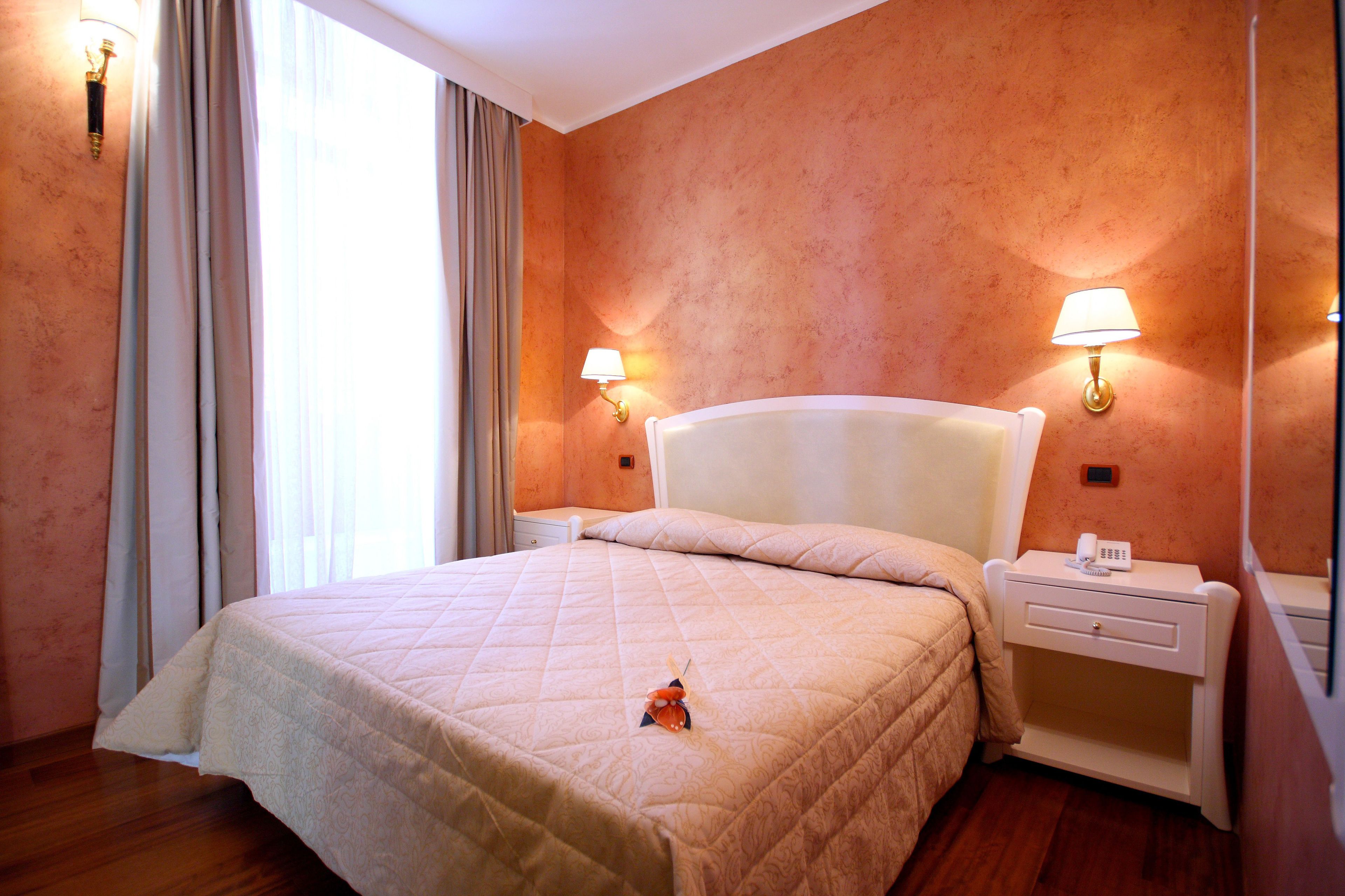 Camera d'albergo con un letto grande, due comodini con lampade, un tavolino con il telefono e una finestra con tende.