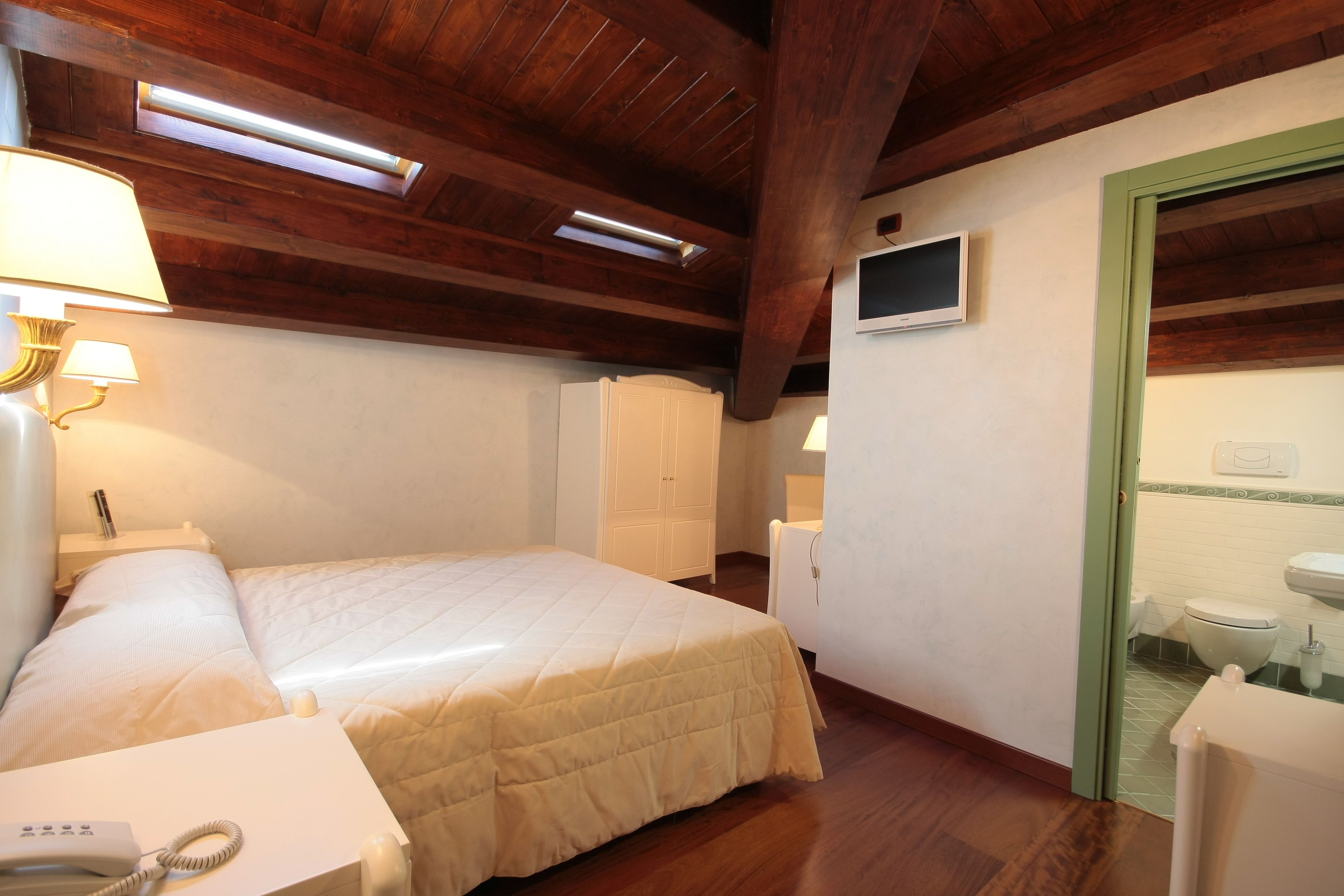 Una camera d'albergo con un letto, lampade sui comodini, una scrivania con un telefono, una TV a parete e un bagno con il water e il lavandino.