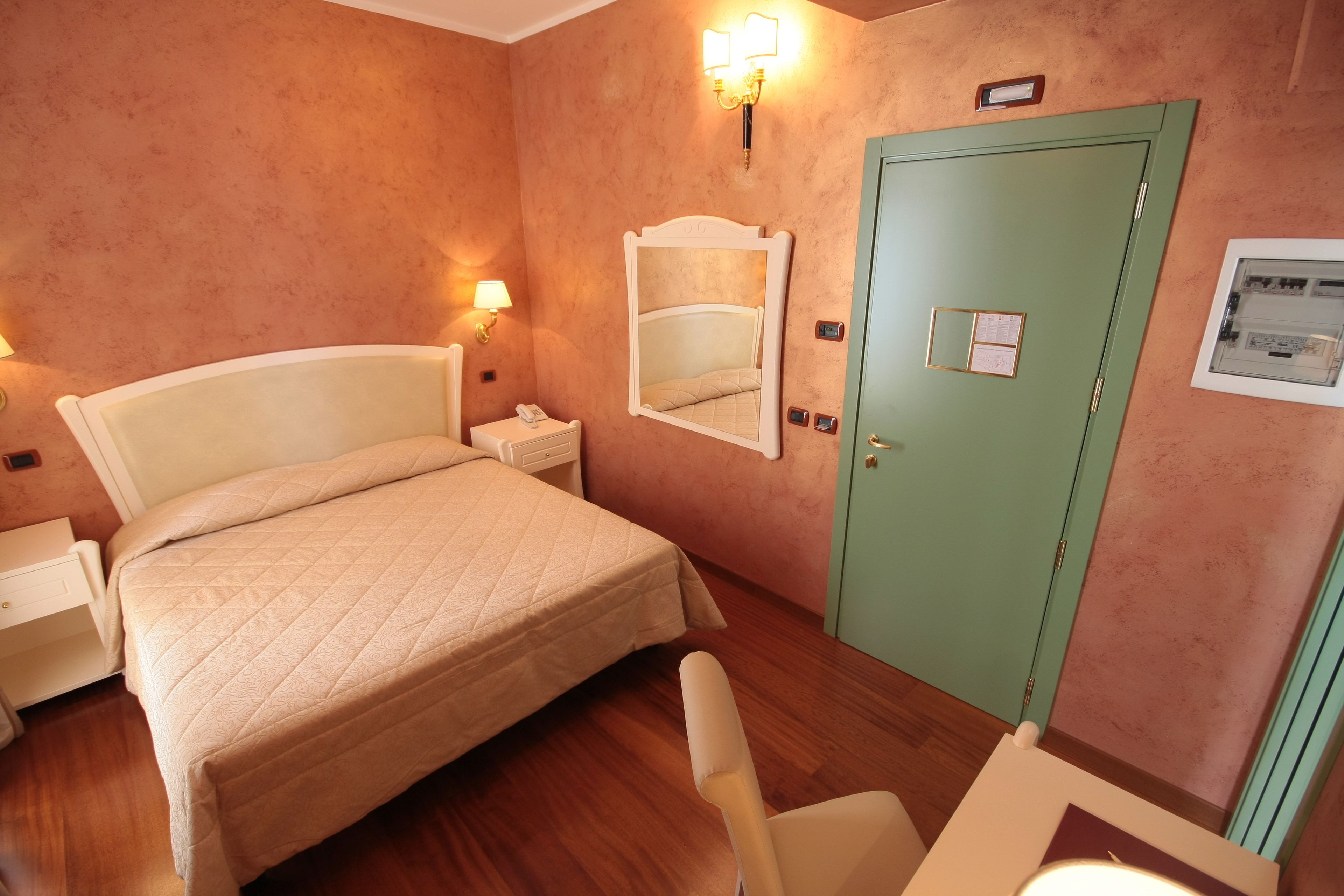 Habitación de hotel con cama, mesitas de noche, un espejo, una puerta verde y un aplique en la pared.