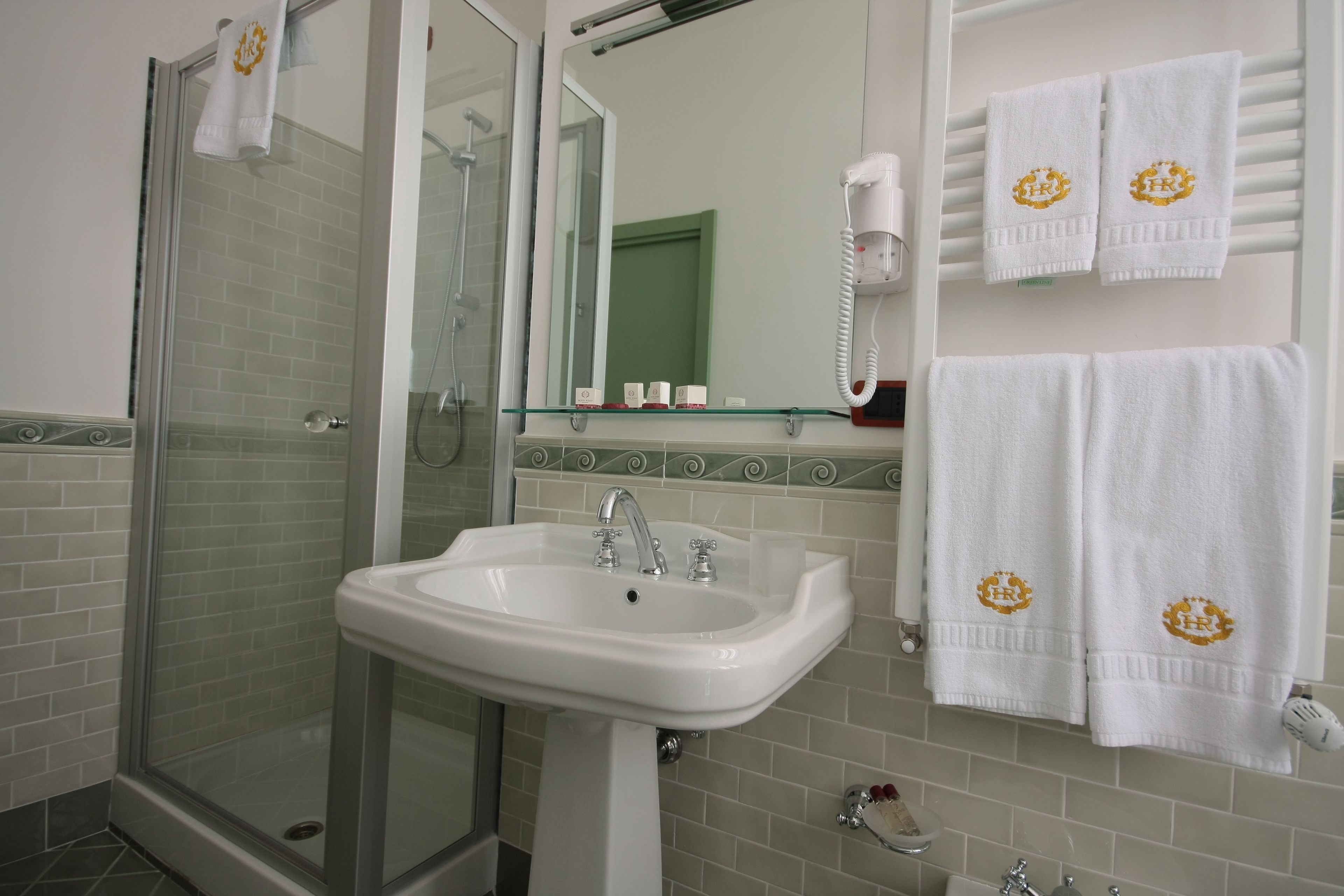 A bathroom with a glass shower enclosure, a white sink with two faucets, and a mirror above the sink. There are two white towels with a logo hanging on a towel rack to the right.