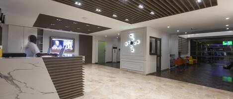 A modern lobby with a reception desk, a large screen displaying a news broadcast, and a seating area with colorful chairs.