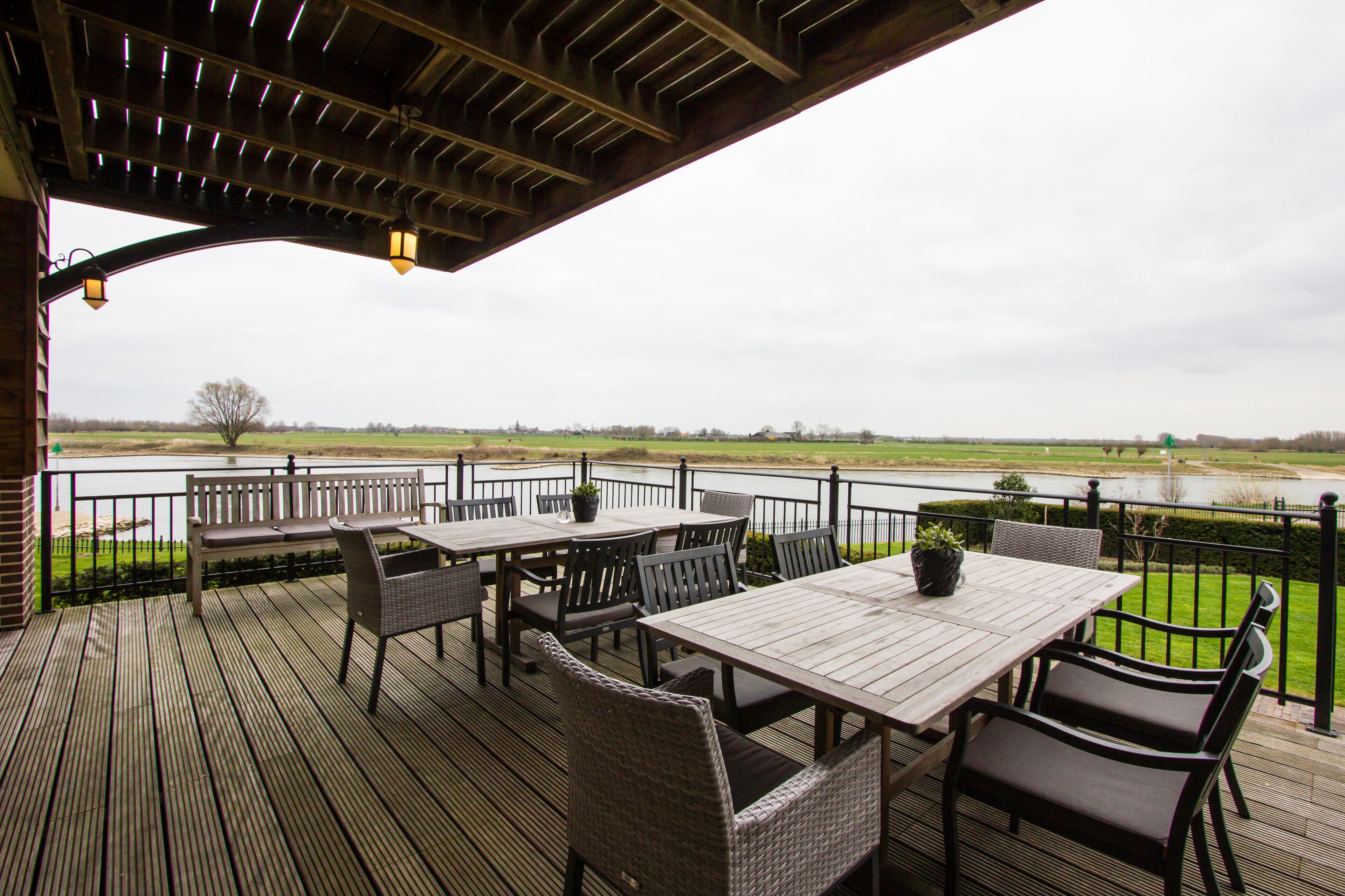 Une terrasse extérieure avec des tables en bois et des chaises en osier, offrant une vue sur une rivière et des champs.