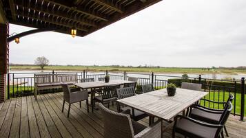 Une terrasse extérieure avec des tables en bois et des chaises en osier, offrant une vue sur une rivière et des champs.