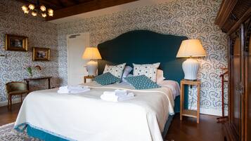 Una cama bien hecha con almohadas y una colcha, una mesita de noche con una lámpara, y una silla en una habitación con papel tapiz estampado y vigas de madera.