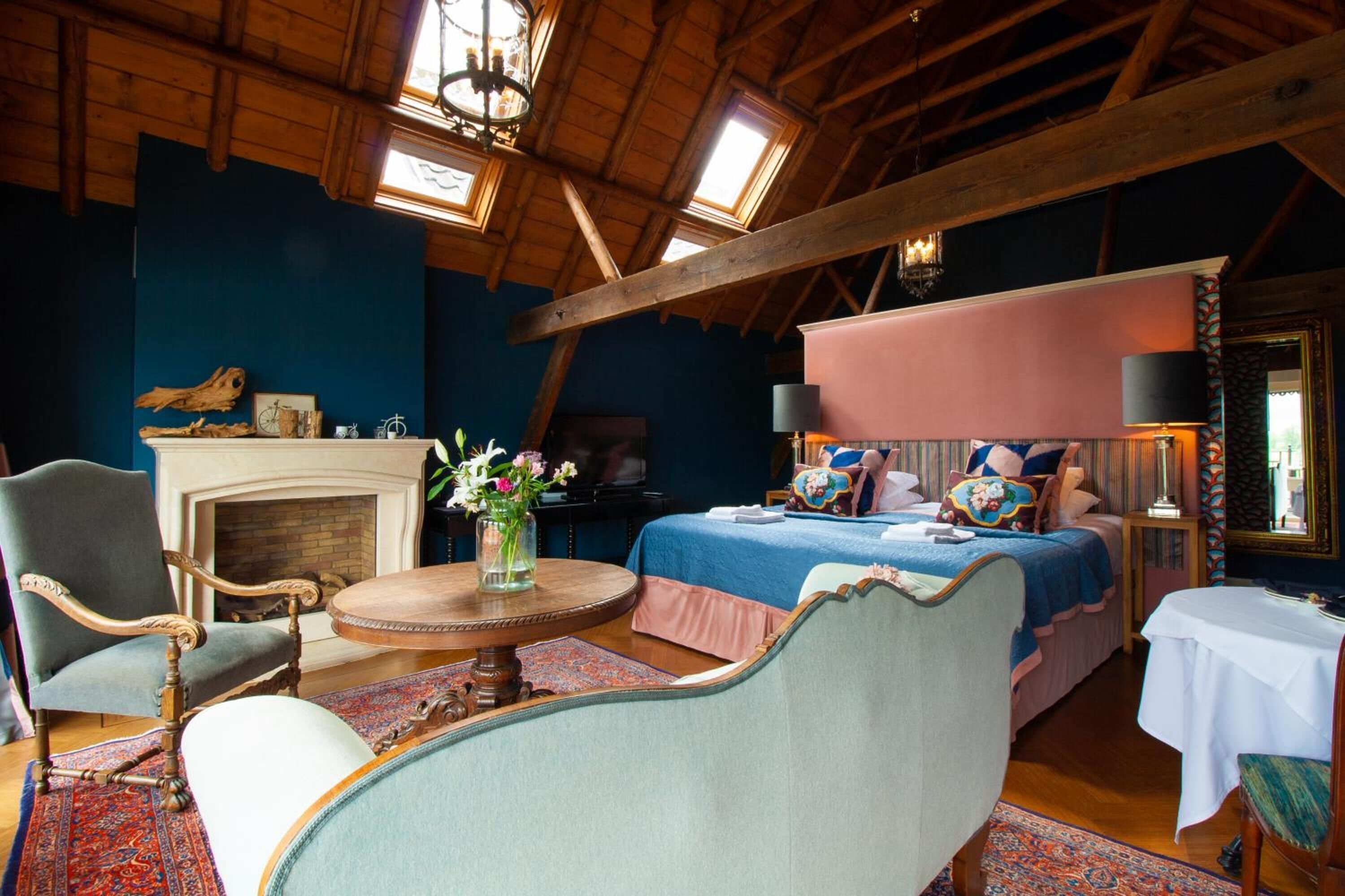 A room with a high ceiling, exposed wooden beams, a fireplace, a bed with a pink canopy, and a round wooden table with a vase of flowers.