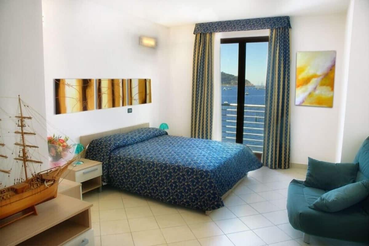 Een slaapkamer met een bed, een nachtkastje, een dressoir, een raam met gordijnen, uitzicht op zee en een schilderij aan de muur.