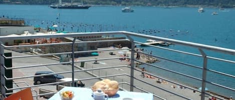 Un balcón con mesa lista para el desayuno, con vista a un puerto deportivo con barcos y una playa.
