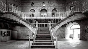 Un grand escalier orné d’une rampe en métal, des murs en briques et des fenêtres en arc de cercle dans un intérieur spacieux.