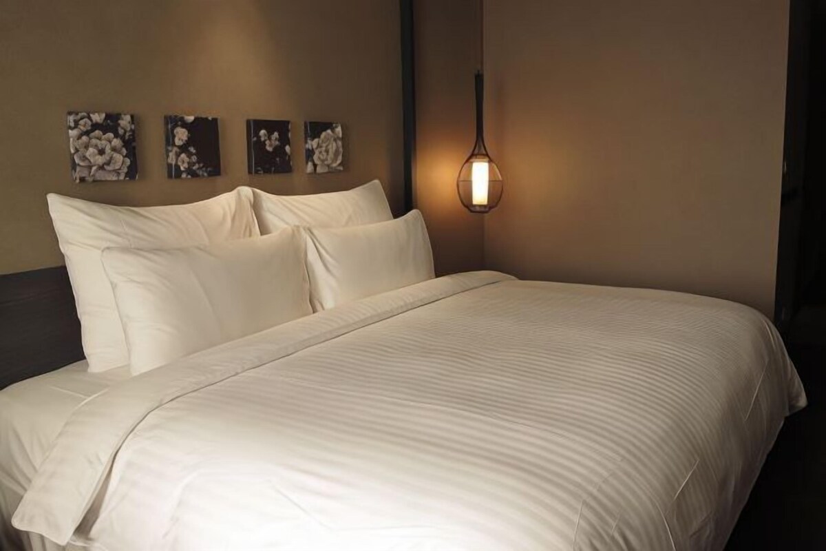 Un lit bien fait, avec du linge de lit blanc, une tête de lit sombre et un luminaire fixé au mur.
