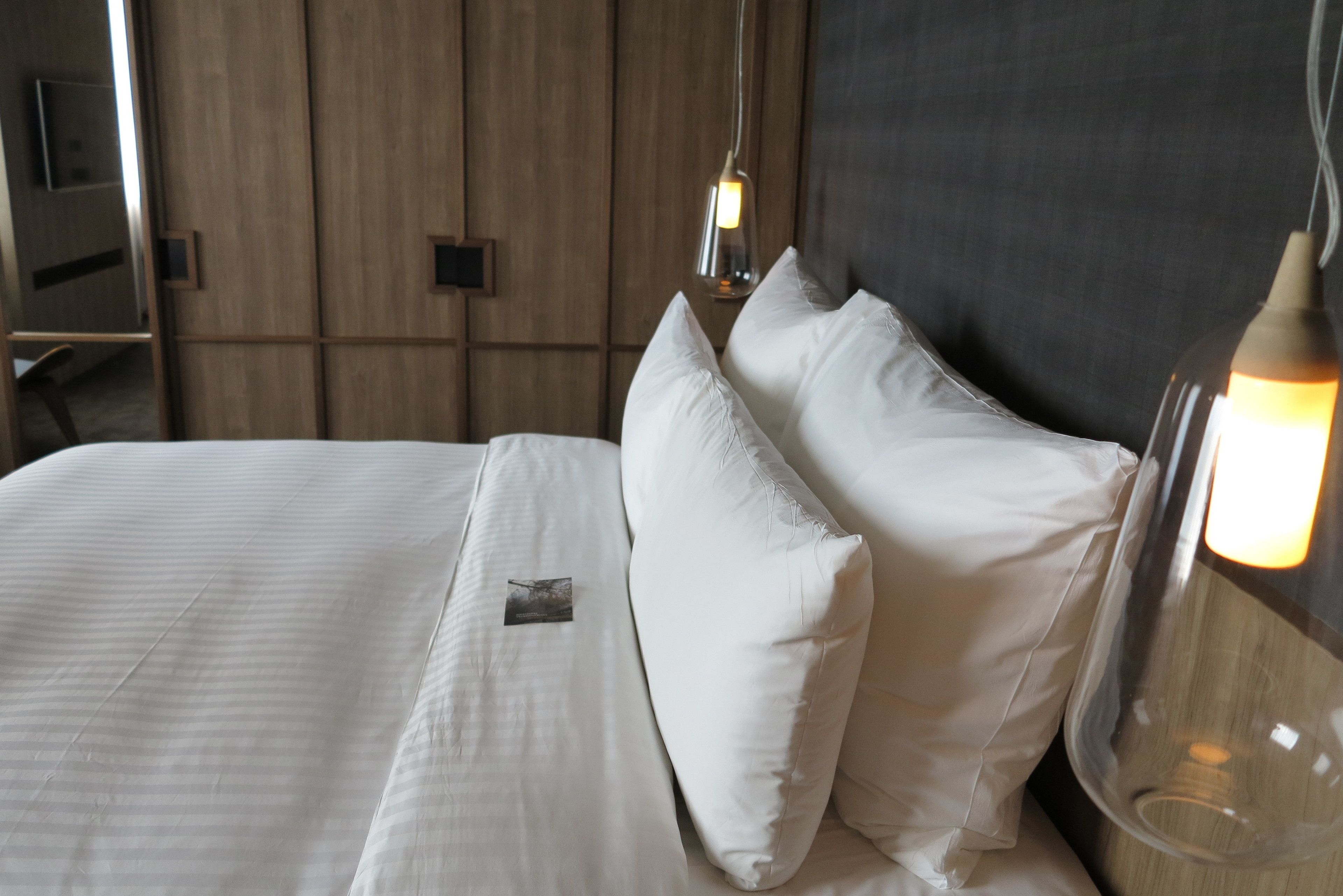 A neatly made bed with white linens and pillows, a bedside table with a lamp, and a wooden panel wall in the background.