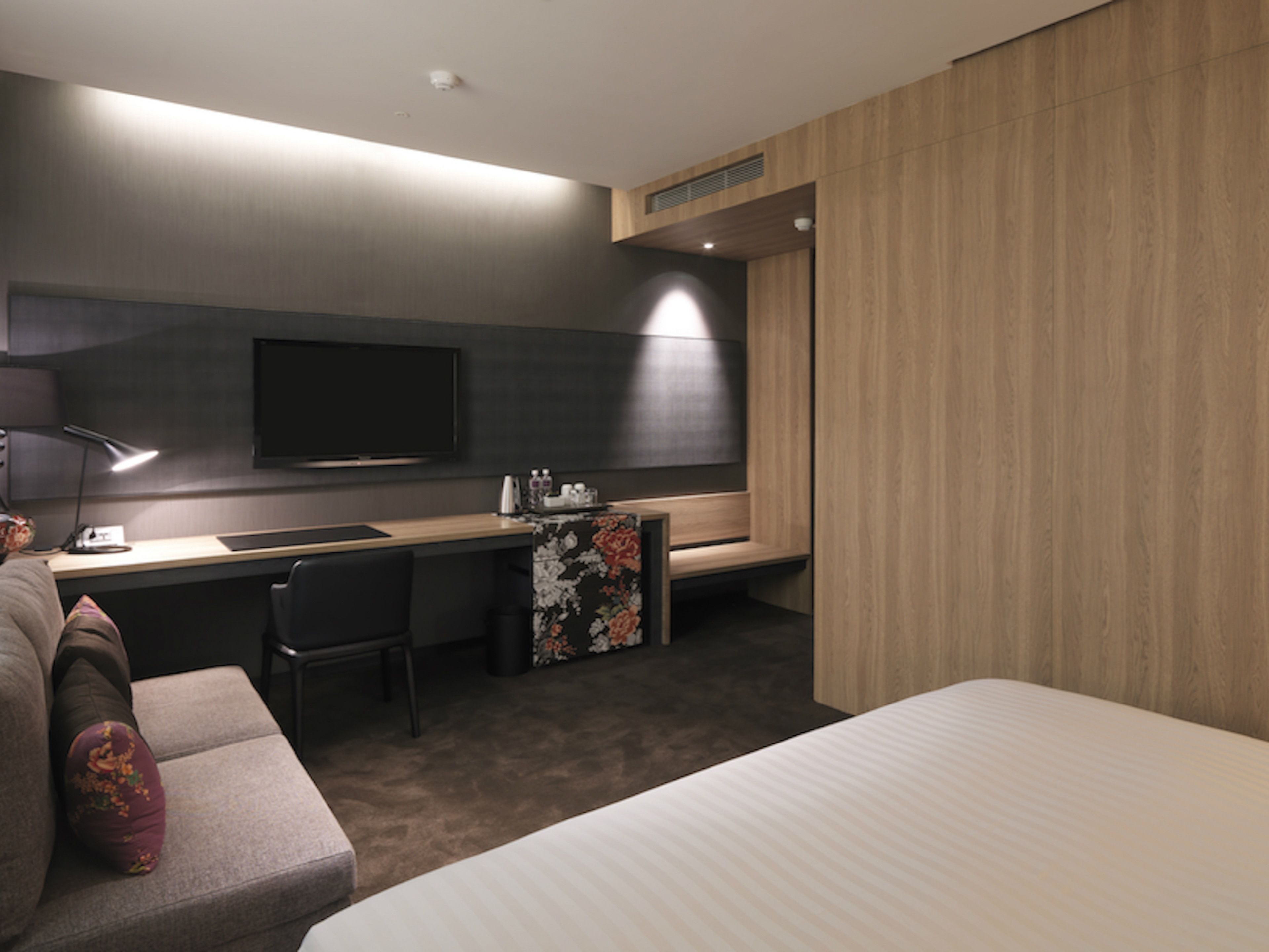 A modern hotel room with a bed, a desk, a chair, a flat-screen TV, and a small table with a floral table runner.