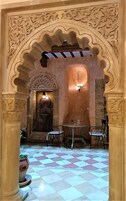 Un interior con piso de tablones, una zona de estar central con sillas y arcos ornamentados.