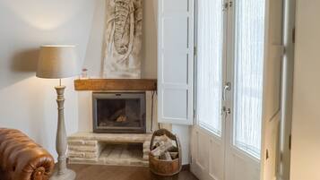 Una sala de estar acogedora con chimenea, vigas de madera, un revestimiento de piedra alrededor de la chimenea, un sofá de cuero y un gran ventanal con contraventanas blancas.