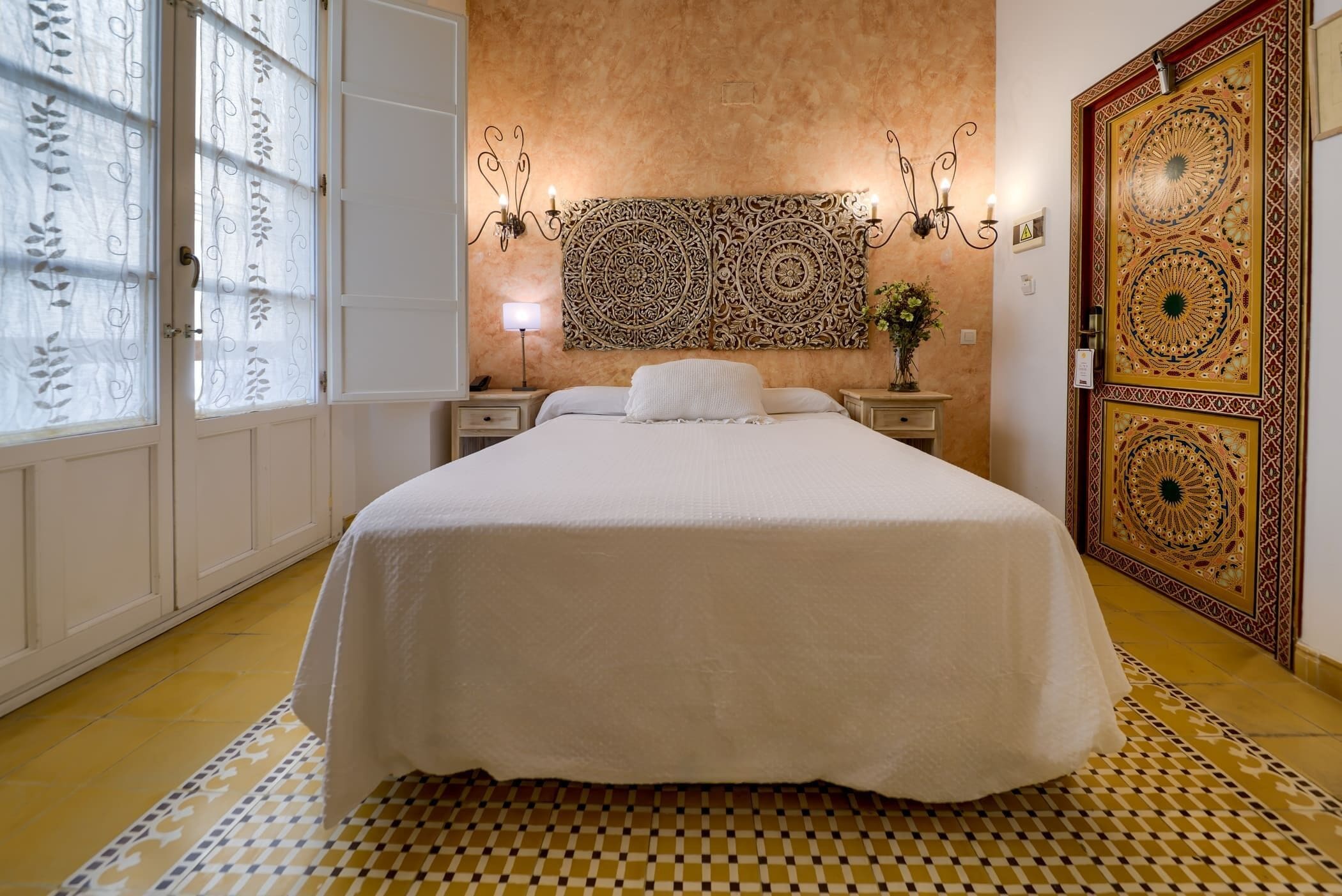A bedroom with a large bed, a patterned rug, a decorative wall piece, and a patterned door.
