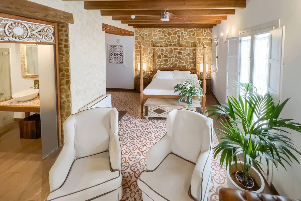A hotel room with a stone wall, a bed with white linens, a wooden ceiling, and a potted plant.