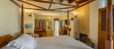 A bedroom with a large bed, wooden ceiling beams, a desk, and a fireplace.