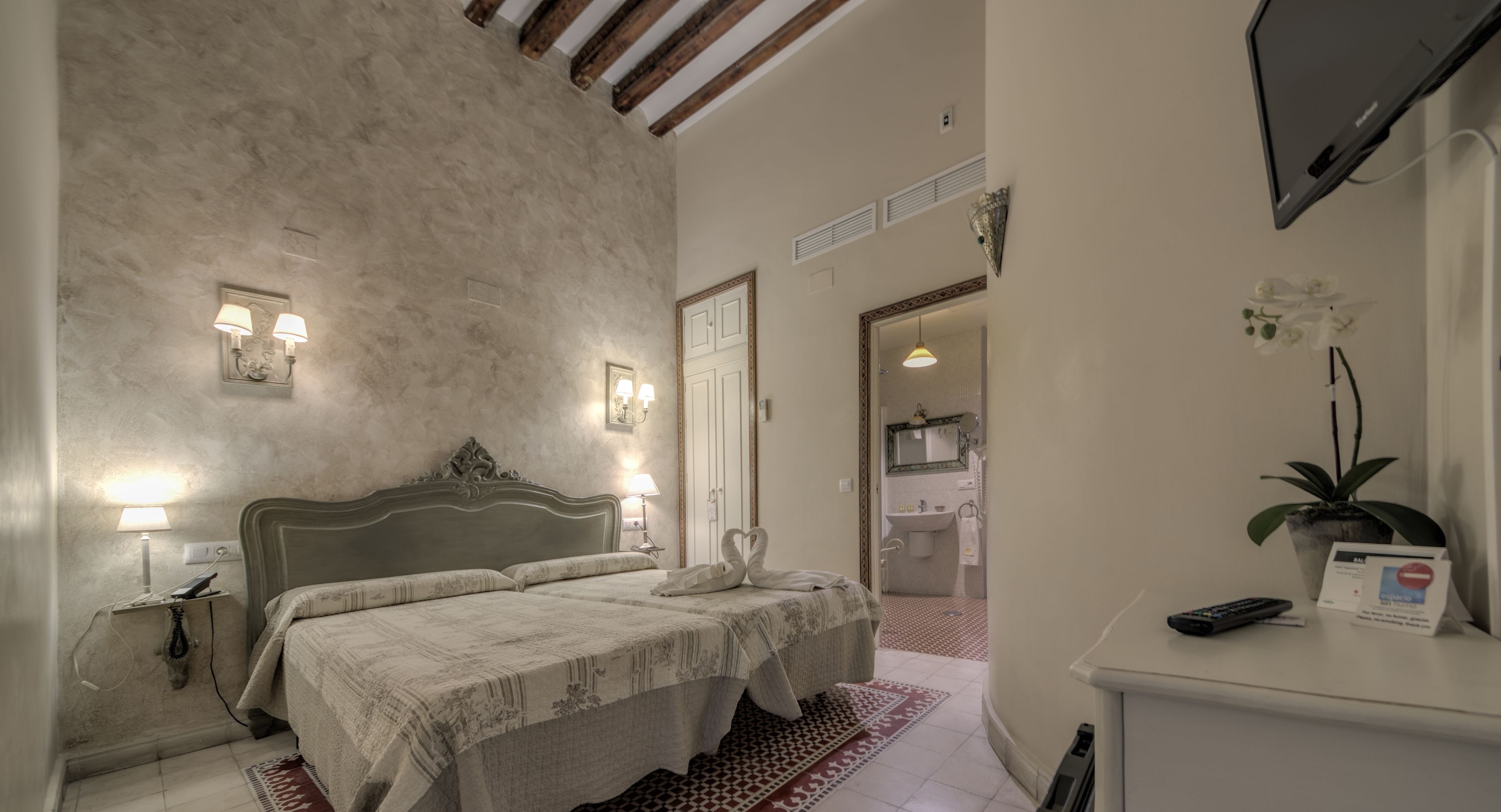 Habitación de hotel con cama, lámparas de noche, escritorio con ordenador y baño visible a través de una puerta abierta.