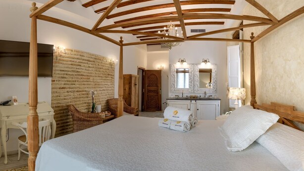 A bedroom with a four-poster bed, a wooden desk, wicker chairs, a brick wall, and a bathroom with a mirror and sink. - Hotel Argantonio (Cádiz)