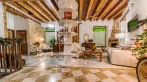 A spacious living room with a checkered floor, a central white bookshelf, wooden beams on the ceiling, a stone wall, and a fireplace. - Hotel Argantonio (Cádiz)