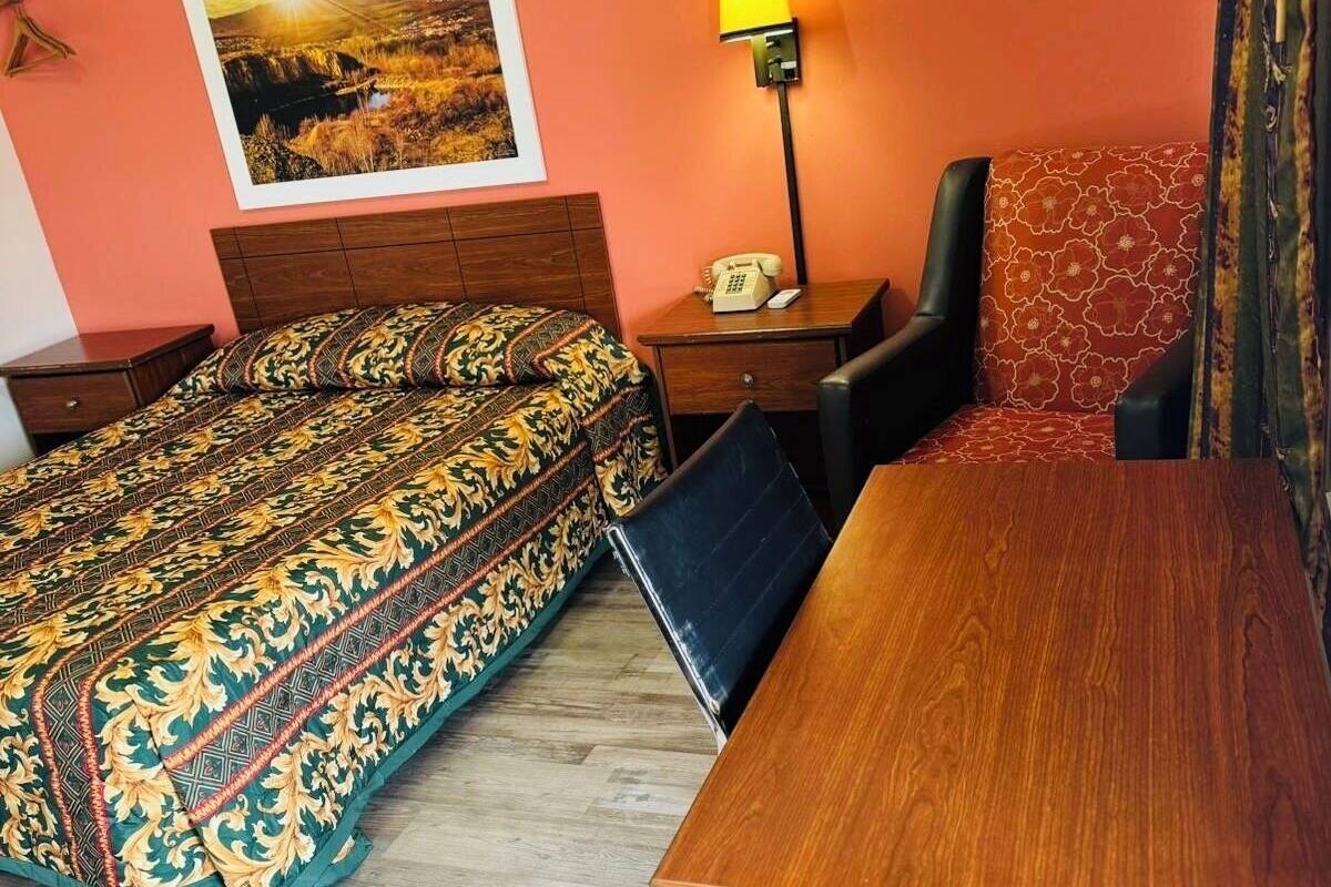 A hotel room with a bed, a desk, a chair, a nightstand, a lamp, and a painting on the wall.
