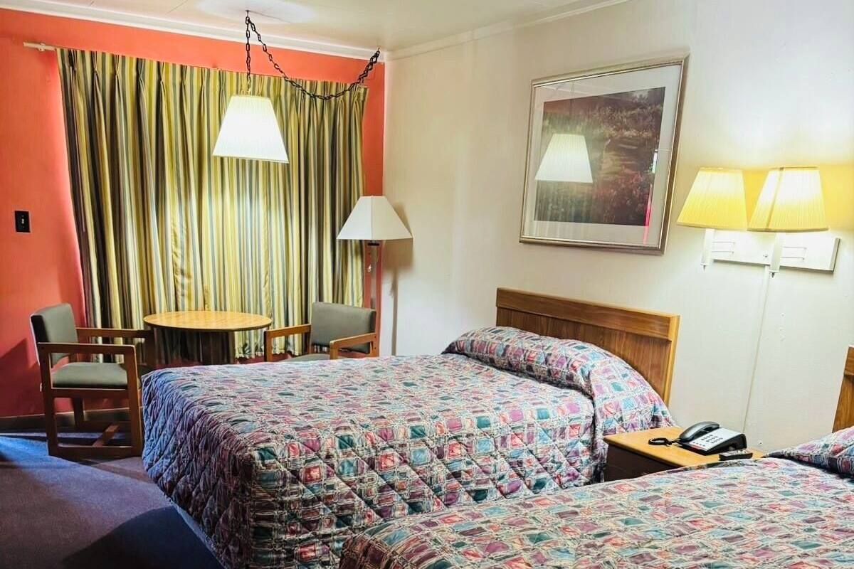 A hotel room with two beds, a table, chairs, a lamp, and a painting on the wall.