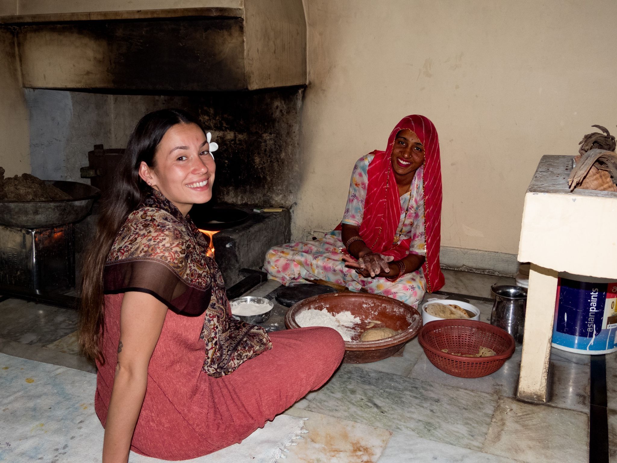 Dos mujeres sentadas en el suelo de una habitación con una estufa grande, tazones y cestas.