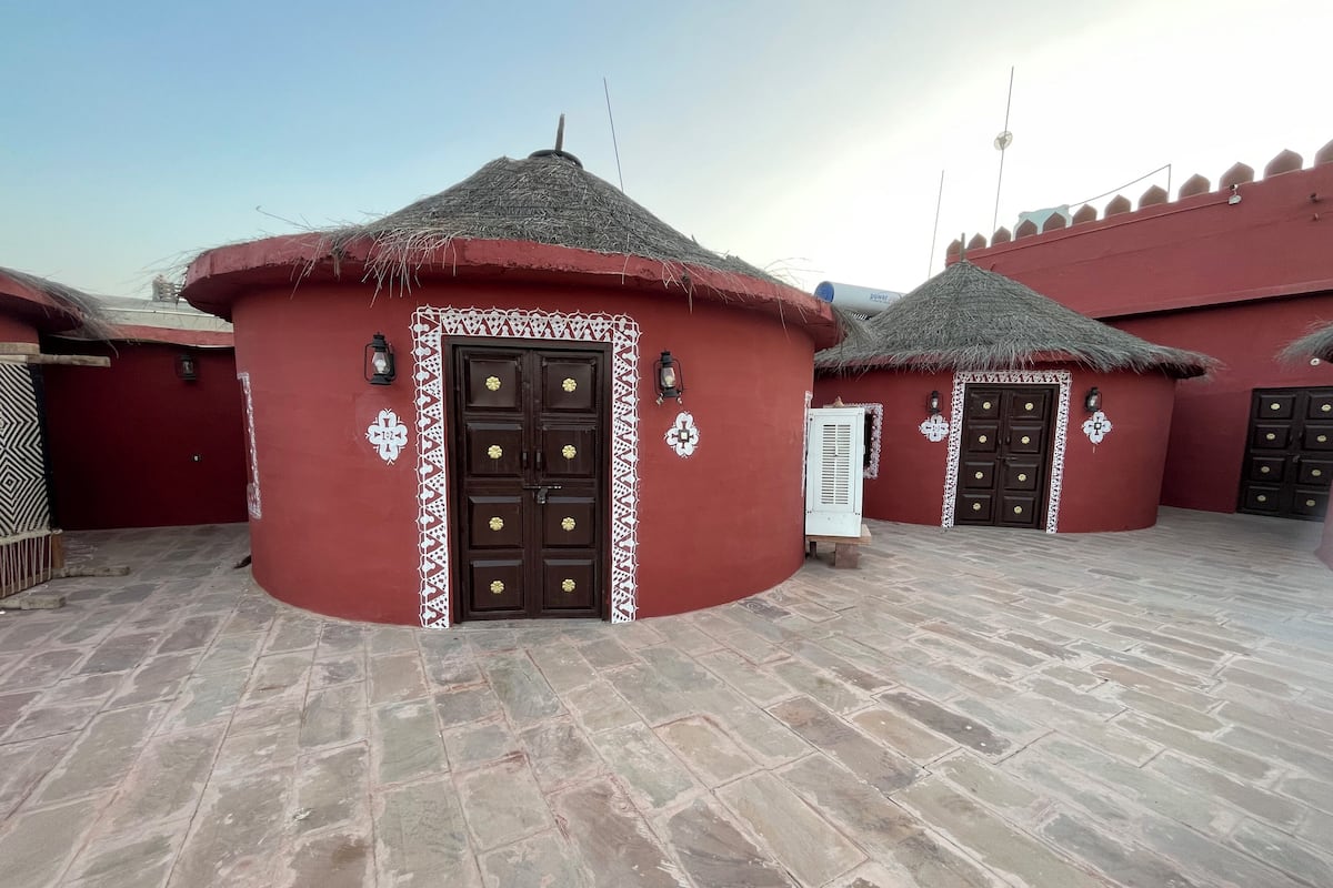 Une cour bordée de bâtiments aux toits rouges, une porte centrale agrémentée d’éléments décoratifs et un sol pavé de pierres.