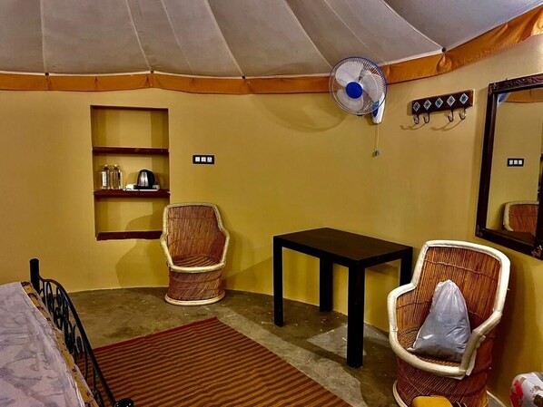 A room with a bed, two wicker chairs, a small table, a fan, a shelf with items, and a mirror on the wall.