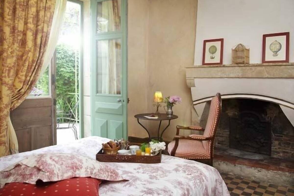 A cozy bedroom with a bed, a fireplace, a chair, and a table with a tray of breakfast items.