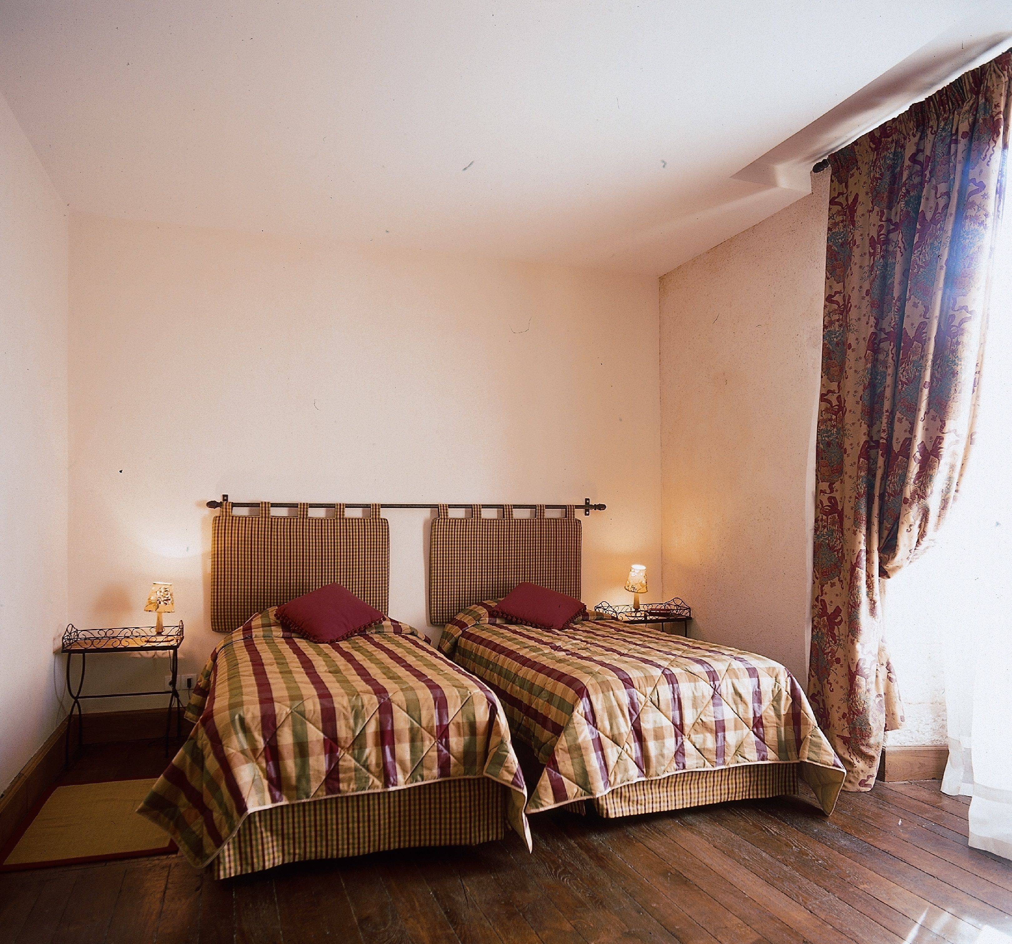 Ein Zimmer mit zwei Betten, jeweils mit kariertem Kopfteil und roten Kissen. An den Betten stehen Nachttische mit Lampen. Der Boden ist mit Holz versehen und ein Fenster ist mit Vorhängen ausgestattet.