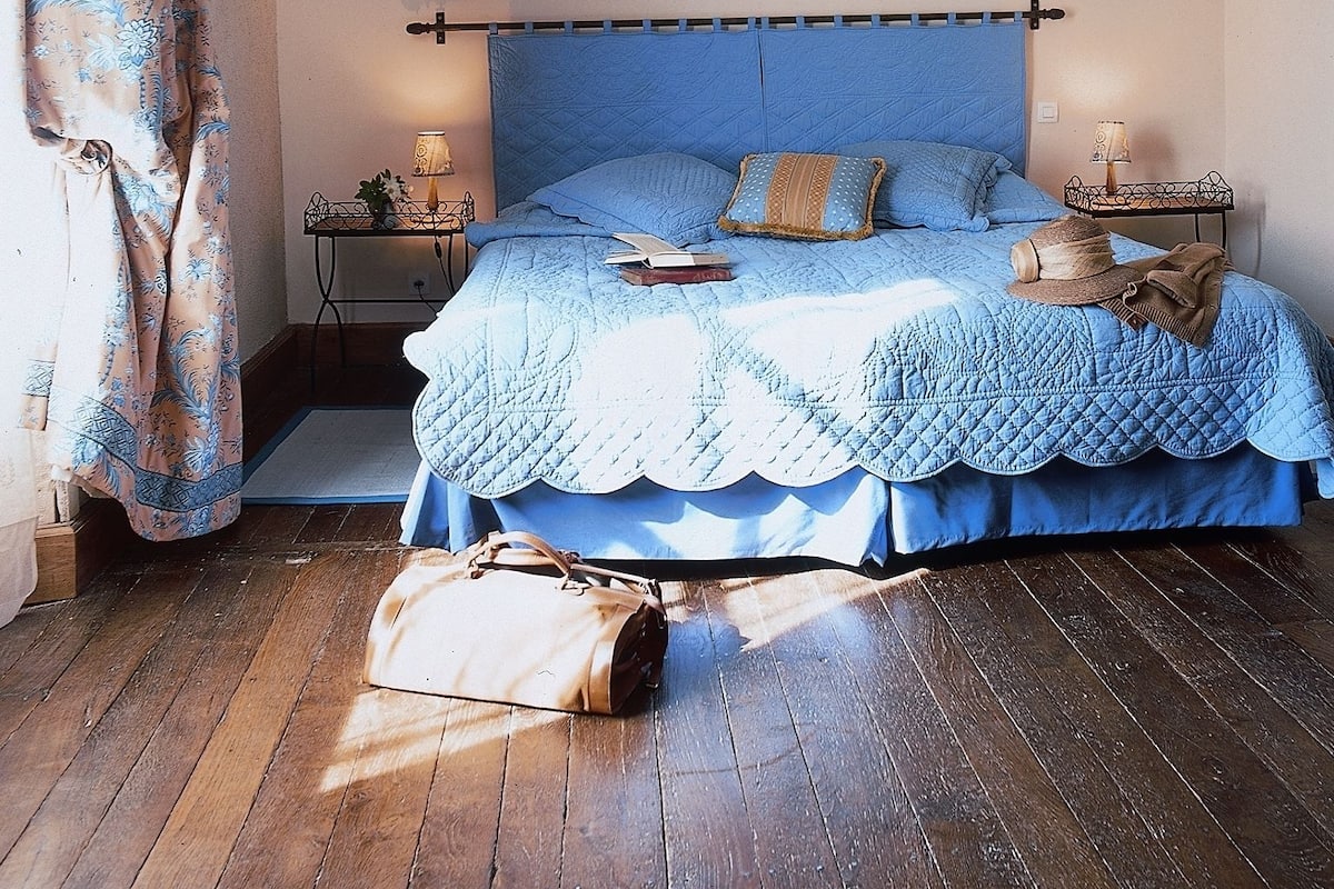 A bedroom with a blue quilted bedspread, a matching blue headboard, and a wooden floor. There are two bedside tables with lamps and a small plant on one. A handbag is on the floor beside the bed.