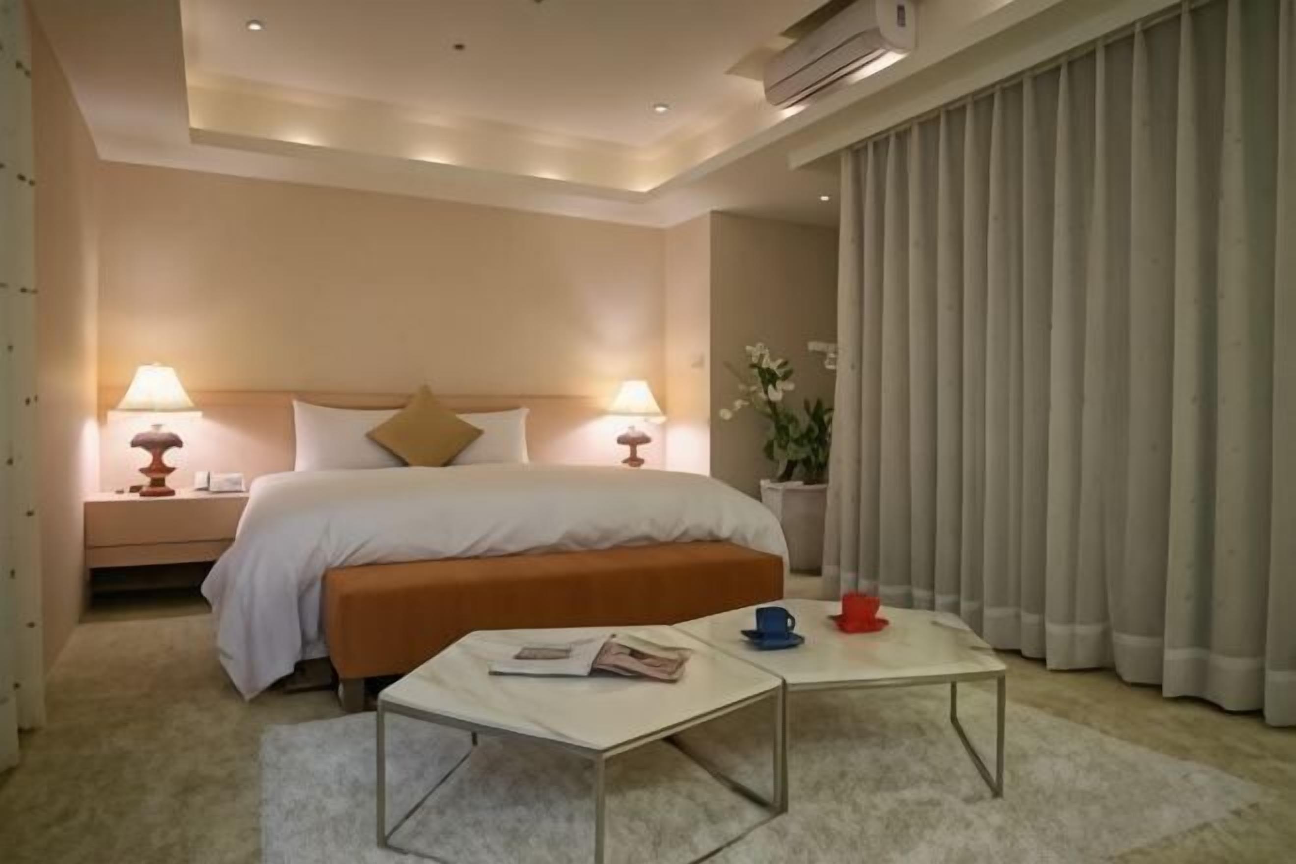 Ein Hotelzimmer mit Bett, Nachttischlampen, einem Nachttisch und einem Couchtisch, sowie einem großen Fenster mit Vorhängen.