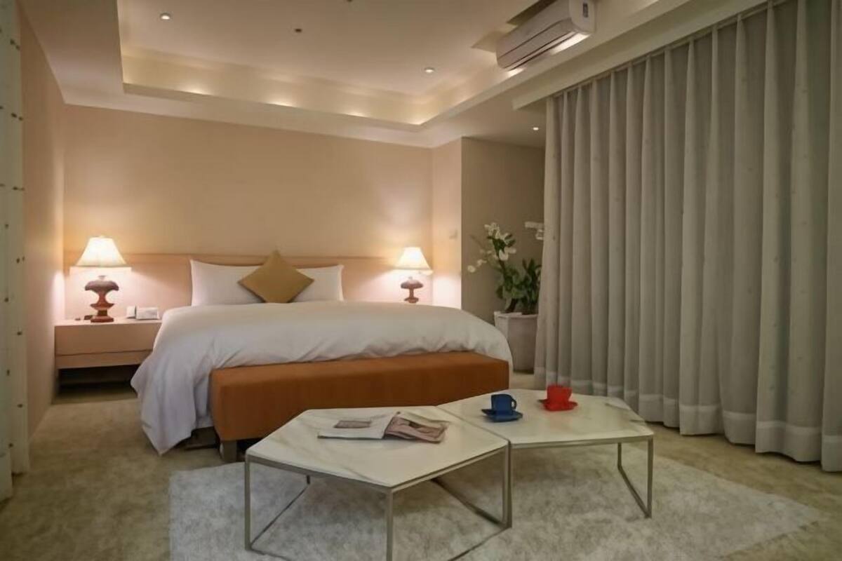A hotel room with a bed, bedside lamps, a nightstand, a coffee table, and a large window with curtains.