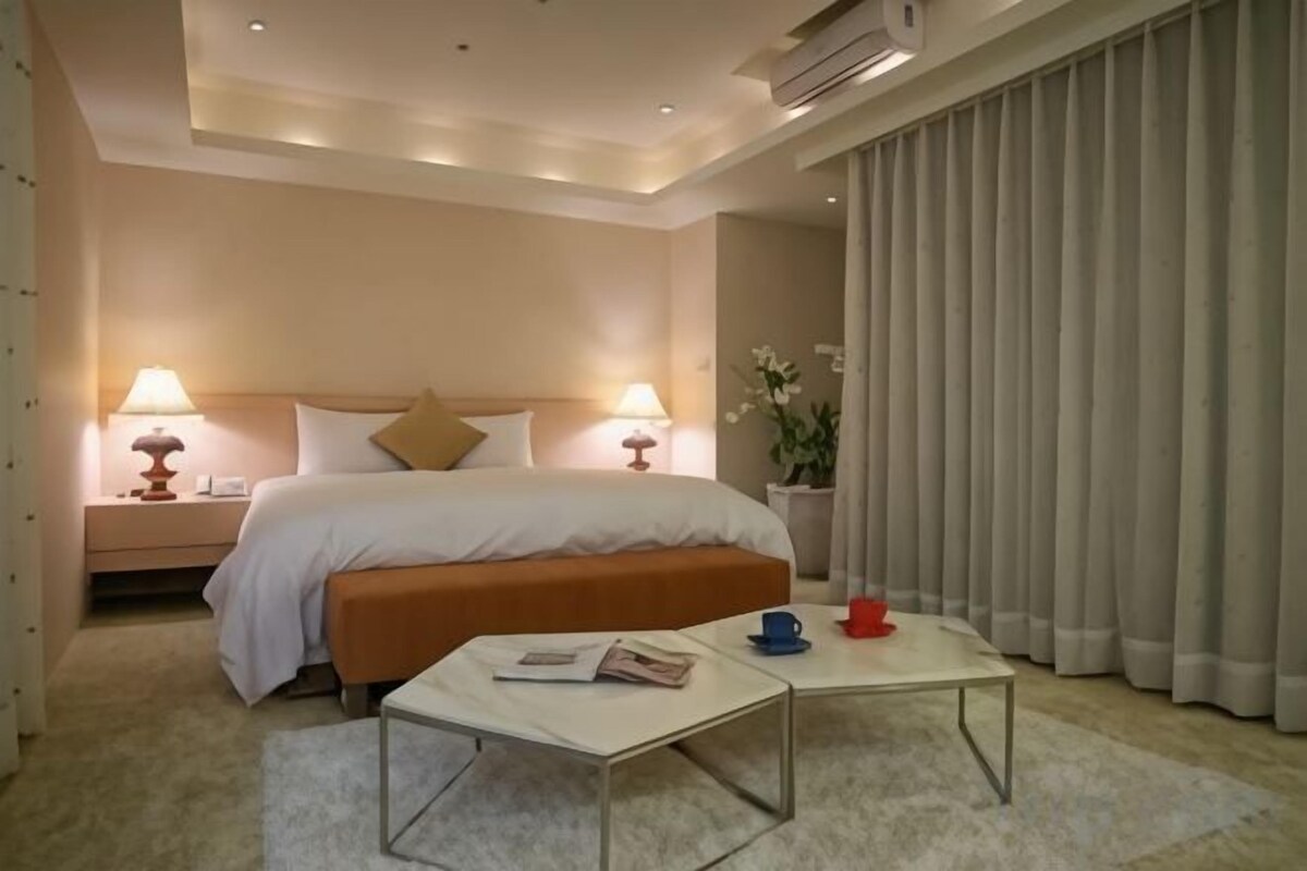 Hotellihuone, jossa on sänky, yöpöytälamput, pieni pöytä ja ikkuna, jossa on verhot.