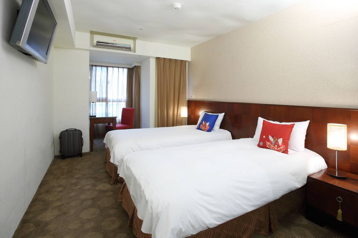 A hotel room with two beds, a television, a lamp, and a suitcase.