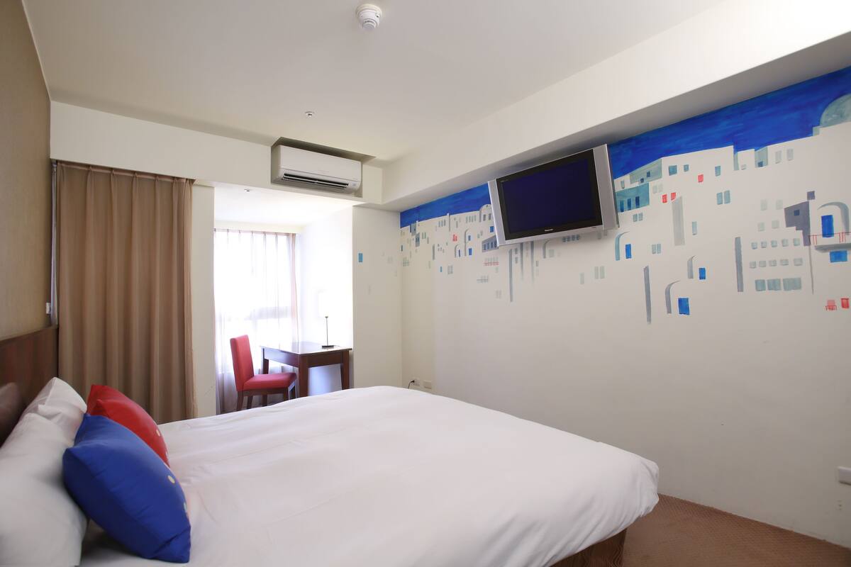 A hotel room with a bed, a TV mounted on the wall, a desk, and a chair.