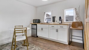 Une cuisine compacte avec des meubles blancs, un réfrigérateur en acier inoxydable, un four à micro-ondes et une machine à café. On y trouve une chaise en bois et une petite table ronde.