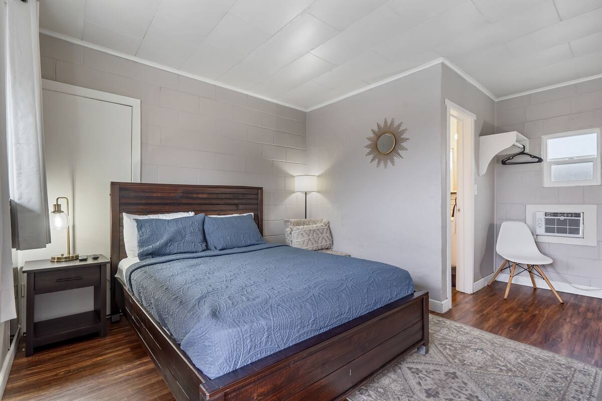 Un dormitorio con una cama de madera, una mesita de noche, una lámpara, una silla y una ventana con aire acondicionado.