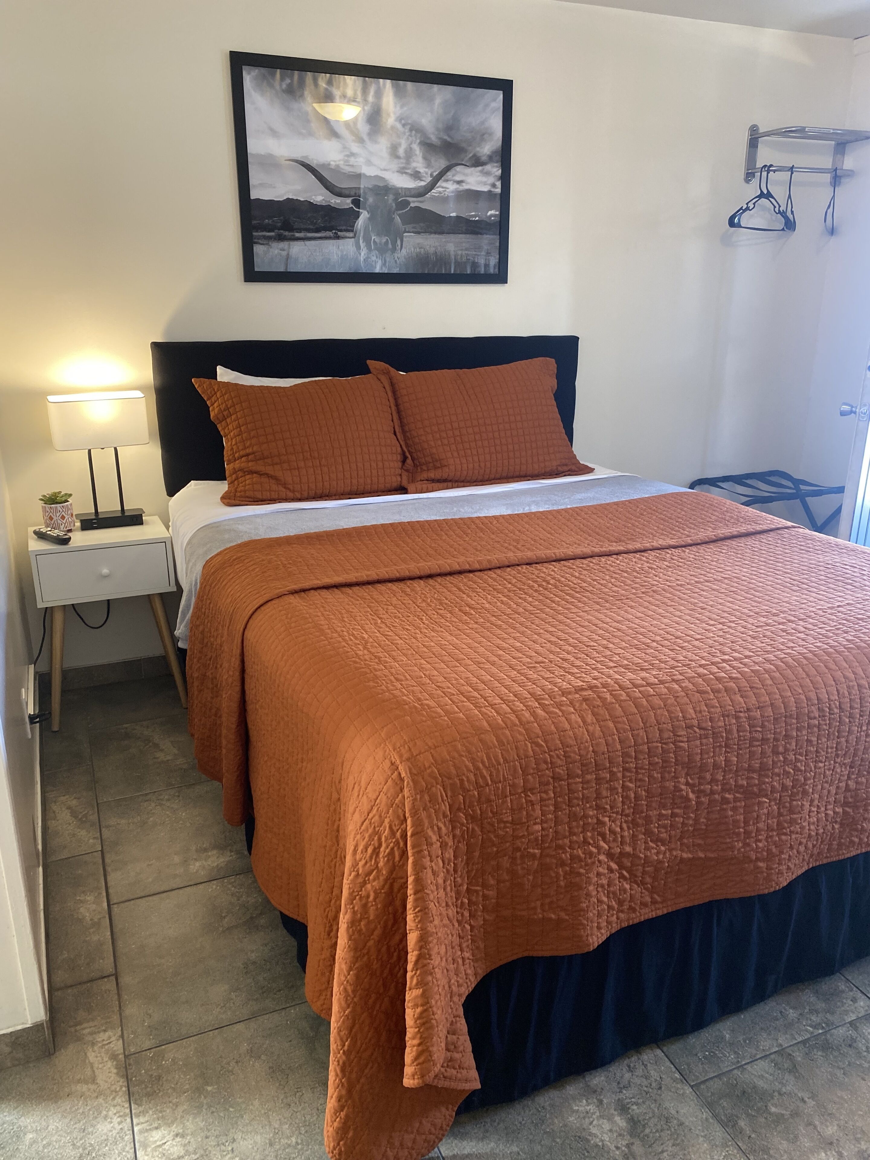 A neatly made bed with an orange quilt, a black headboard, and a framed picture above it.
