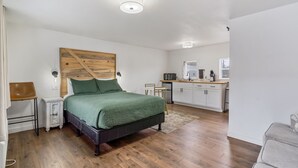 Une chambre avec une tête de lit en bois, une couvre-lit verte, une table de chevet, un bureau et, au fond, un coin cuisine.