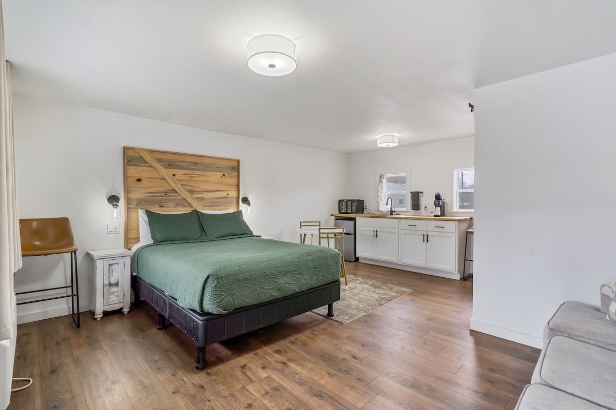 Un dormitorio con cabecera de madera, colcha verde, mesita de noche, escritorio y una kitchenette al fondo.