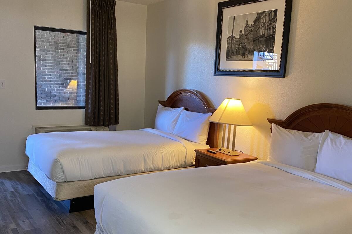 A hotel room with two beds, a nightstand with a lamp, a framed picture on the wall, and a window with curtains.