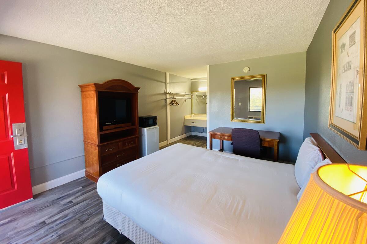 A hotel room with a large bed, a desk with a chair, a television, a small refrigerator, a red door, and a bathroom with a shower and a mirror.