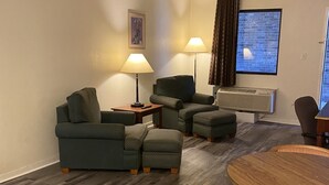 Una sala de estar con un sofá, un reposapiés y una mesa redonda con sillas. Hay dos lámparas, un cuadro en la pared y una ventana con cortinas.